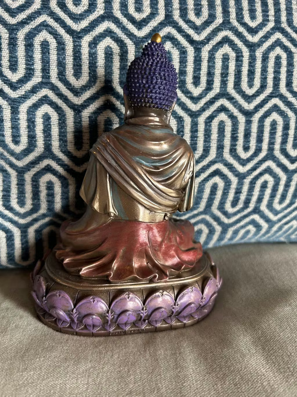 The back of the Buddha statue with purple hair and base against a patterned fabric background