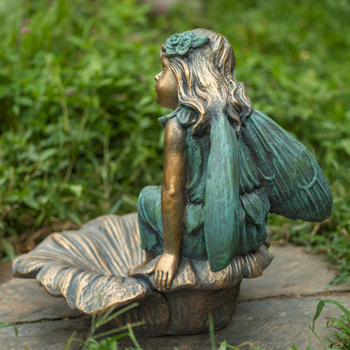 Bronze fairy statue with green wings and hair sitting on a stone surface with grass in the background