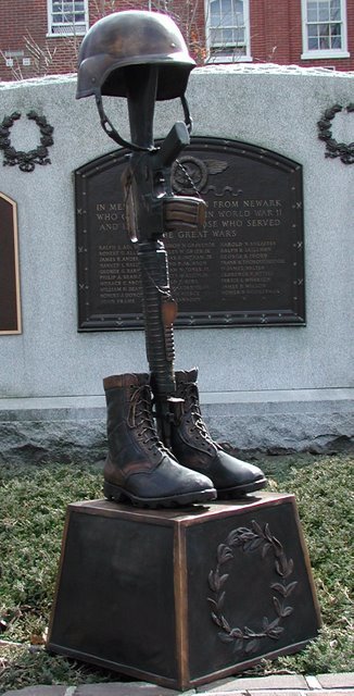Memorial sculpture with helmet, boots, and rifle on a pedestal in front of a wall with inscriptions.