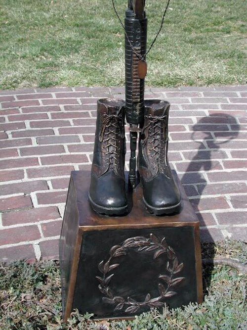 Bronze sculpture of boots and a rifle on a pedestal with a wreath emblem, set against a brick pavement and grass background.