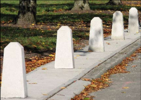 A row of white concrete post caps or bollards installed along a sidewalk next to a road with scattered autumn leaves, with grass and tree trunks in the background.