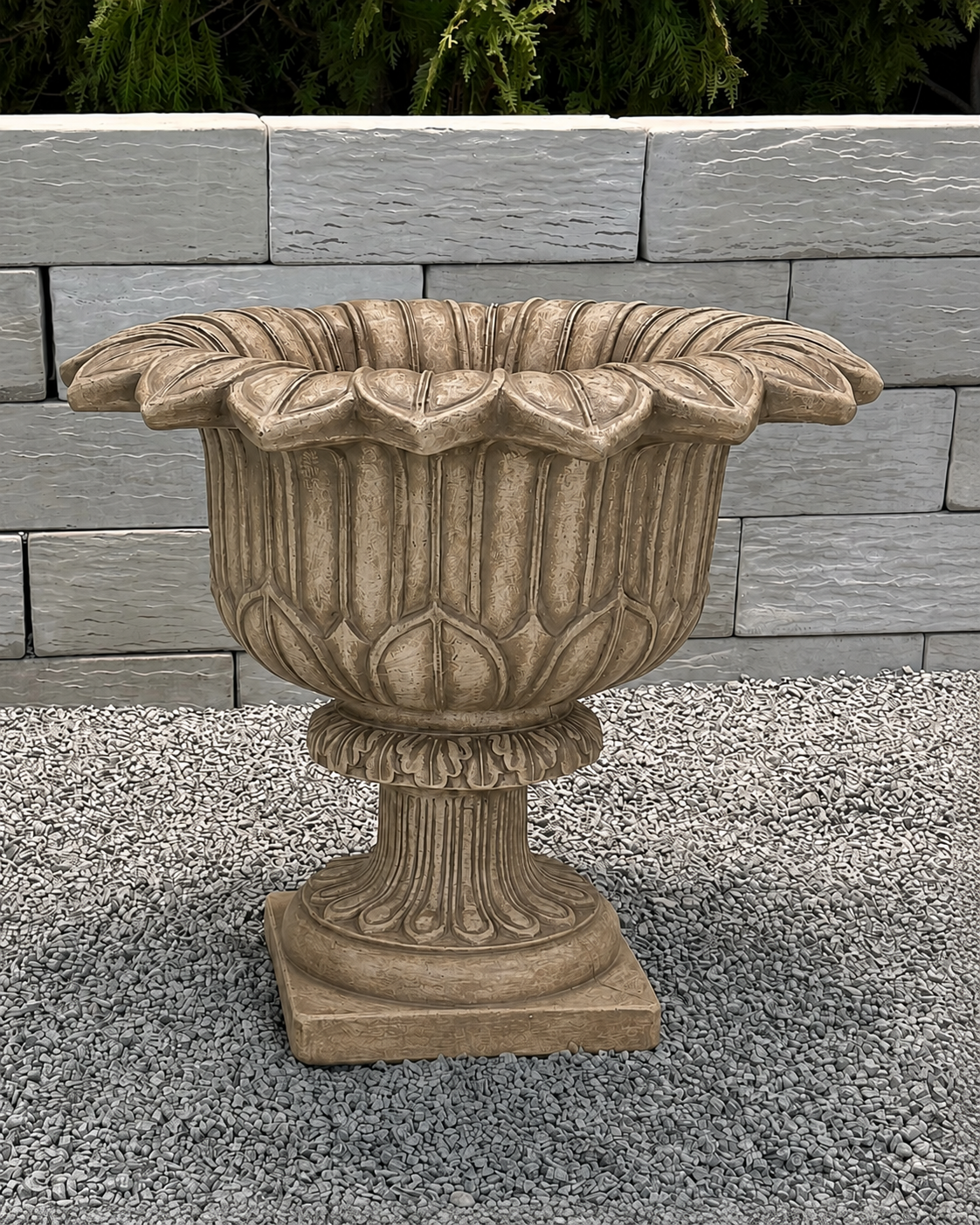 An ornate stone-look planter pot with a fluted pedestal base and wide, scalloped rim, shown outdoors on gravel in front of a light gray stone wall.