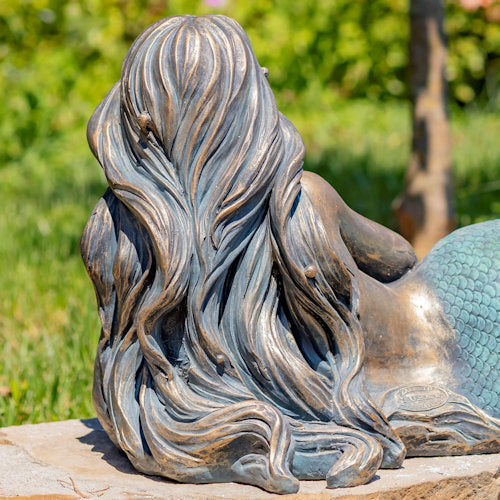 Bronze mermaid statue with flowing hair and scales, set against a natural background.