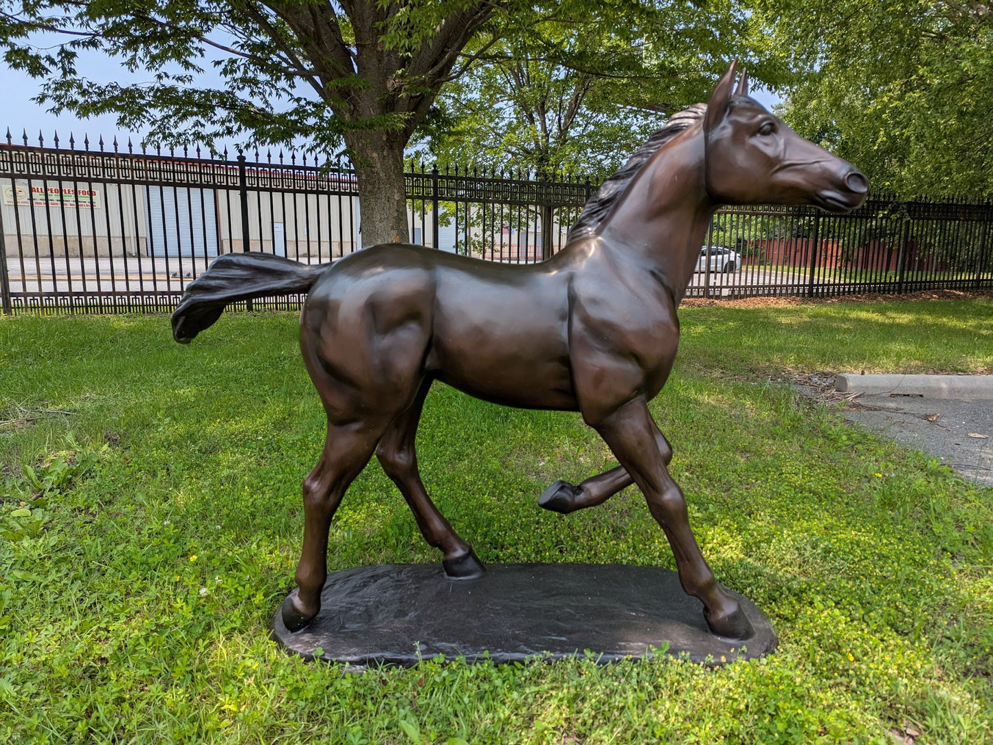 Bronze statue of a horse on grass with a fence and trees in the background