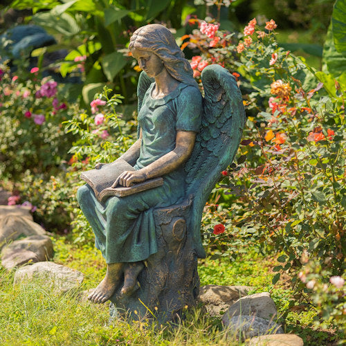Statue of an angel reading a book in a garden setting with flowers and plants.