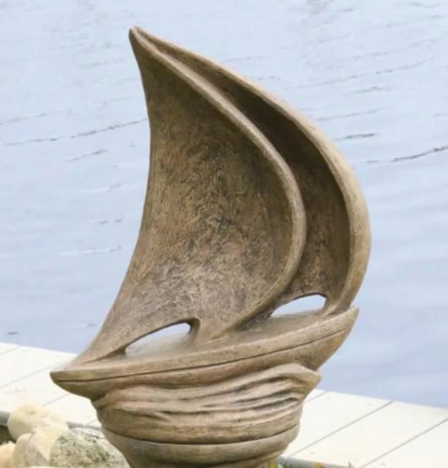 A sculptural garden ornament shaped like a stylized sailboat with a textured stone-like finish, placed outdoors near water and a light-colored dock or deck.