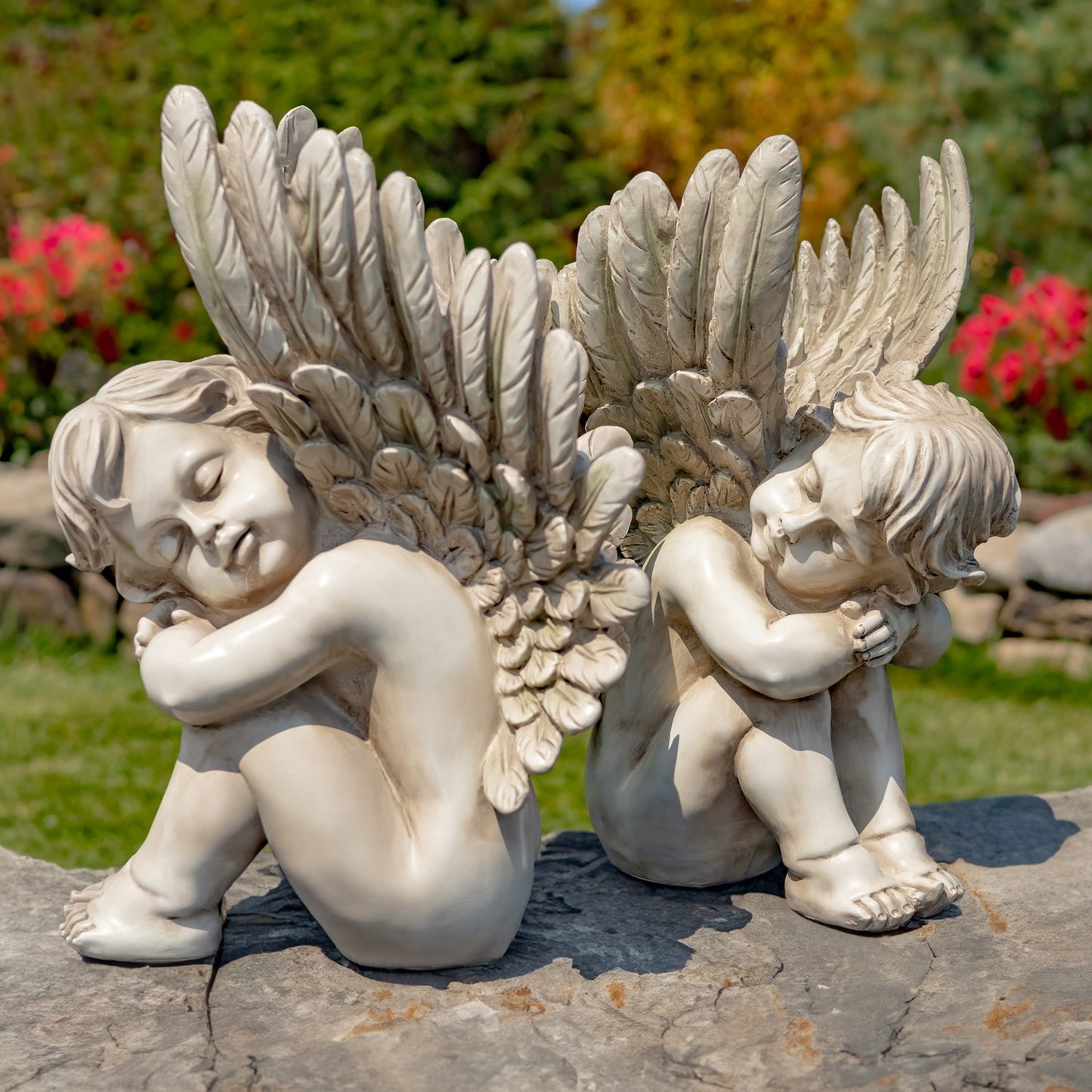 Two angel garden statues with large feathered wings, shown seated on a stone surface outdoors with grass, trees, and flowers in the background.