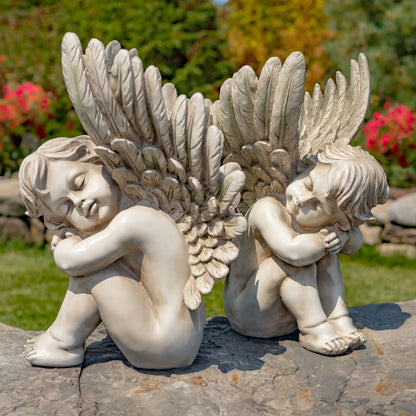 Two angel garden statues with large feathered wings, shown seated on a stone surface outdoors with grass, trees, and flowers in the background.
