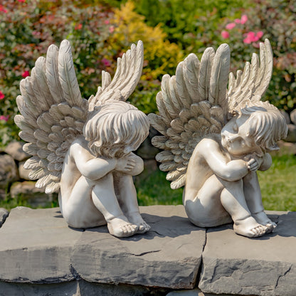 Two white angel garden statues with large wings and crouching, hugging poses, displayed on a stone wall in a garden setting.