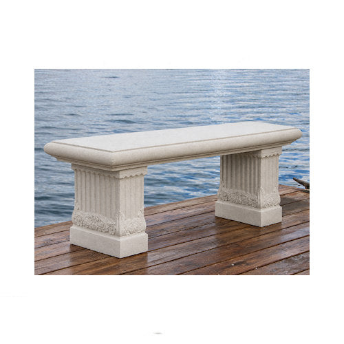 A rectangular outdoor bench made of light beige stone or concrete, featuring fluted column-style supports, shown on a wooden dock with water in the background.