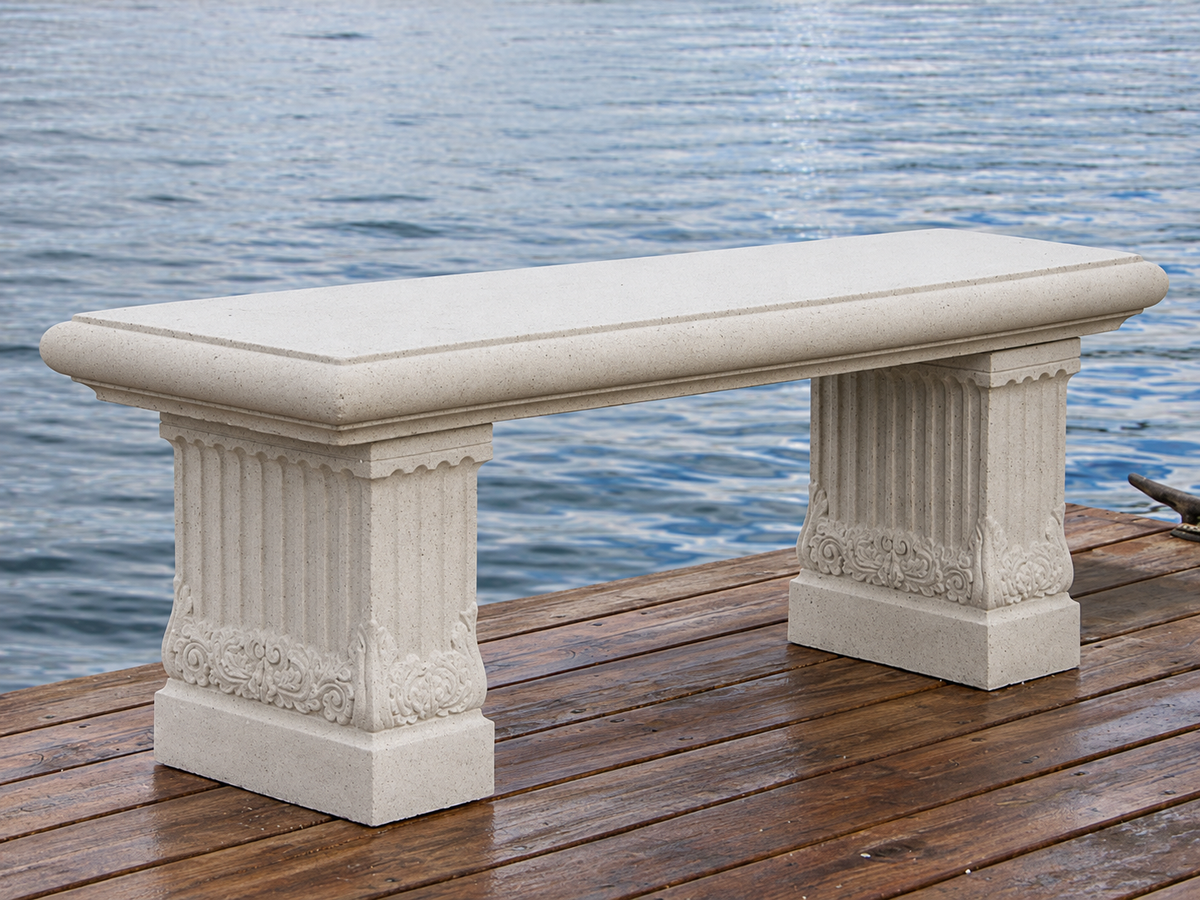 A rectangular outdoor bench made of light beige stone, featuring fluted column-style legs and decorative carvings, positioned on a wooden dock with water in the background.