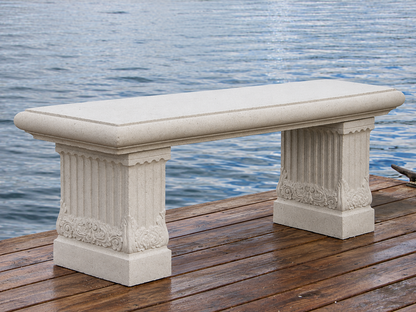 A rectangular outdoor bench made of light beige stone, featuring fluted column-style legs and decorative carvings, positioned on a wooden dock with water in the background.