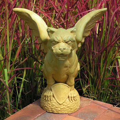 A winged gargoyle garden statue in a light olive-green color, perched on a textured base with a shield emblem, set against a background of tall red and green ornamental grass.