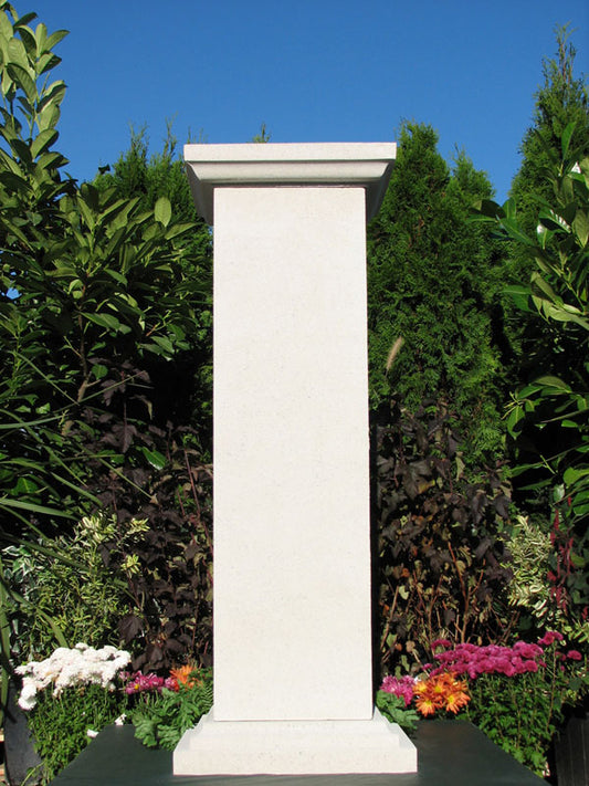A tall, rectangular white stone or concrete monument pillar standing outdoors in a garden setting with green foliage, flowers, and a blue sky background.