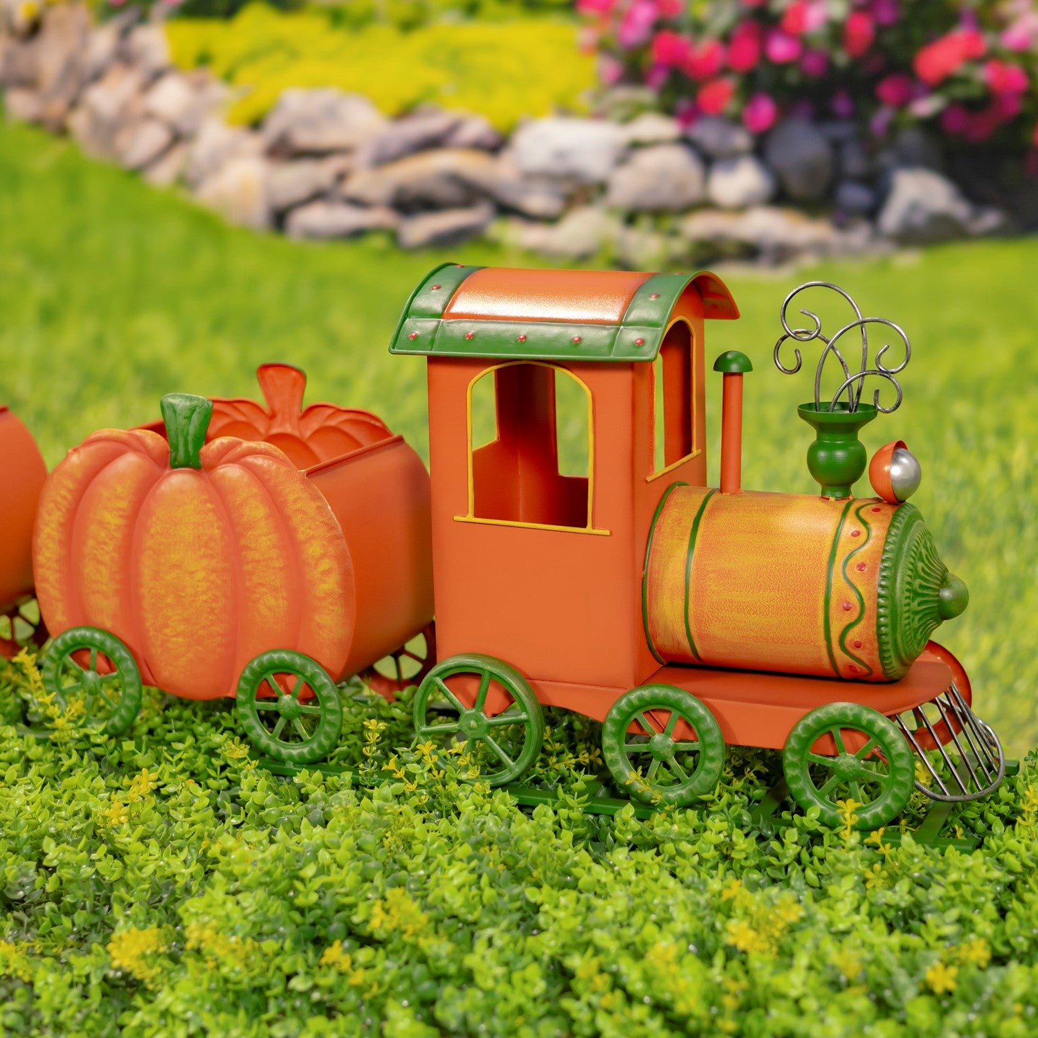 A decorative metal garden train shaped like a pumpkin car and locomotive, displayed outdoors on artificial green grass with blurred flowers and rocks in the background.