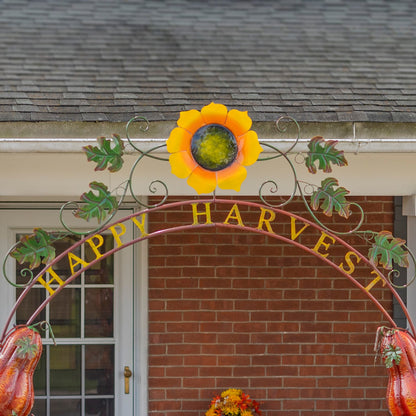 A decorative metal autumn arch with a large yellow sunflower at the center, green leaves, hanging gourds, and the words “HAPPY HARVEST” in gold letters, set against a brick house facade.
