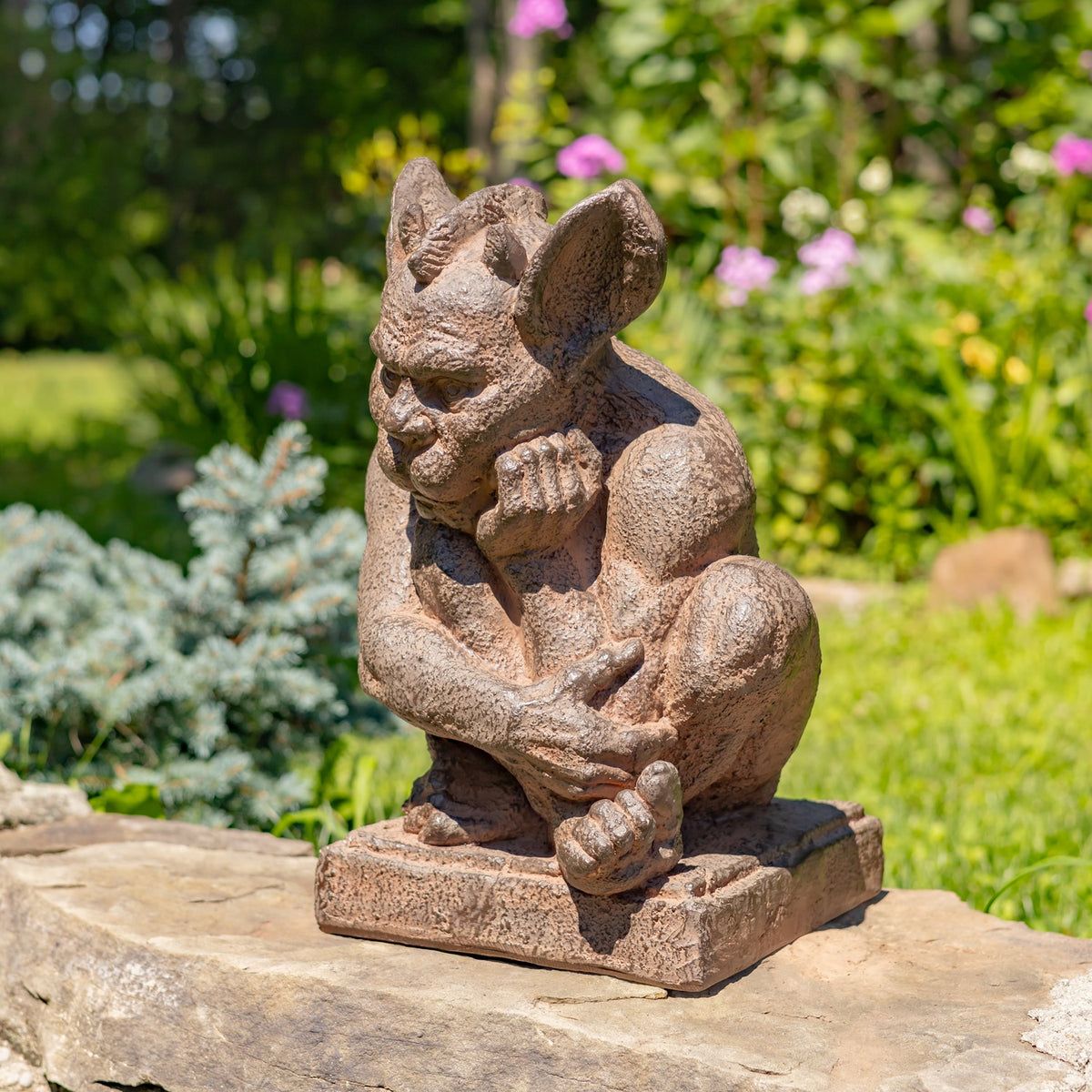 A small stone gargoyle garden statue in a crouched pose, placed on a large stone slab in a lush green garden with blurred plants and flowers in the background.