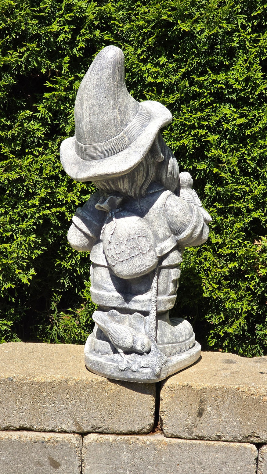 A gray stone garden gnome figurine standing on a stone wall with a dense green hedge in the background.