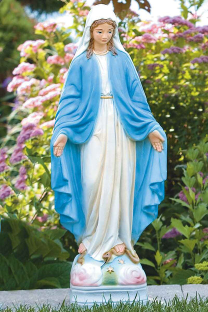 A painted Virgin Mary garden statue with a blue cloak and white gown, standing outdoors in front of a blurred garden background of green foliage and pink flowers.