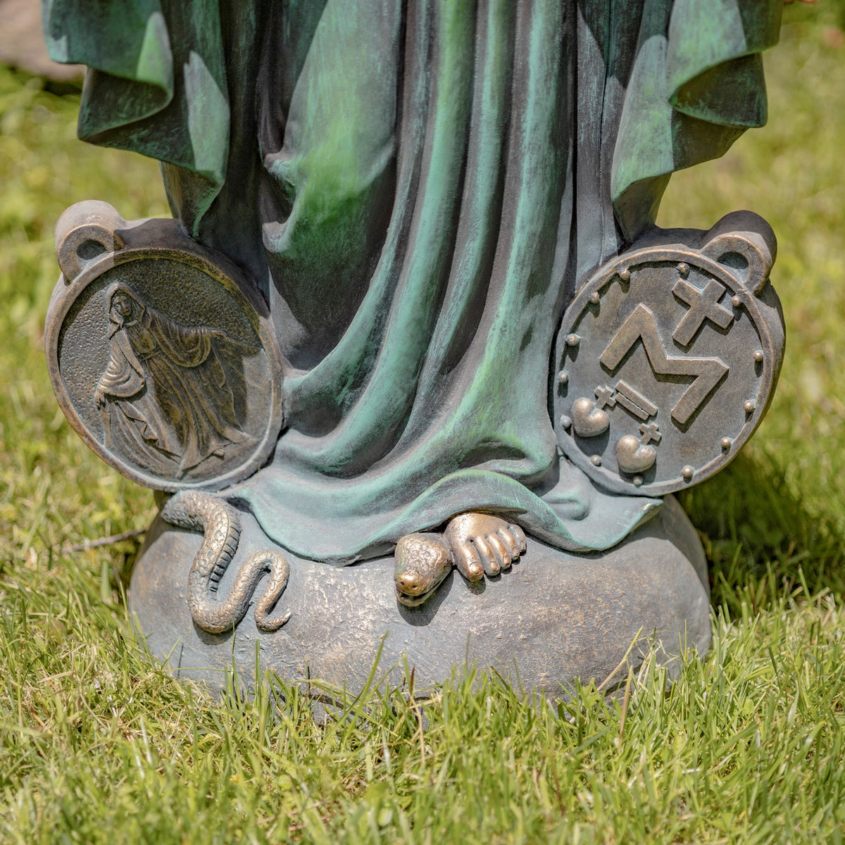 Close-up of a decorative statue base outdoors on grass, showing draped greenish-gray fabric, a snake at the left, a small foot at the center, and two circular metal medallions on either side.