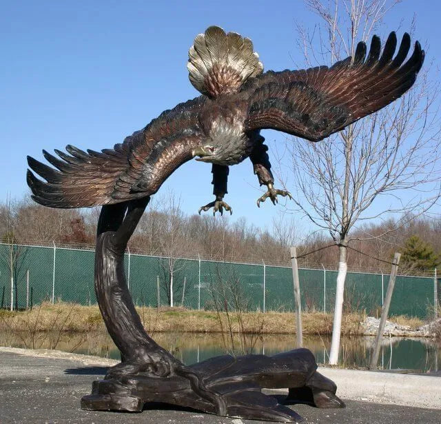 A large bronze eagle sculpture with wings spread wide, perched on a curved tree trunk base, photographed outdoors near a fence, trees, and water under a blue sky.