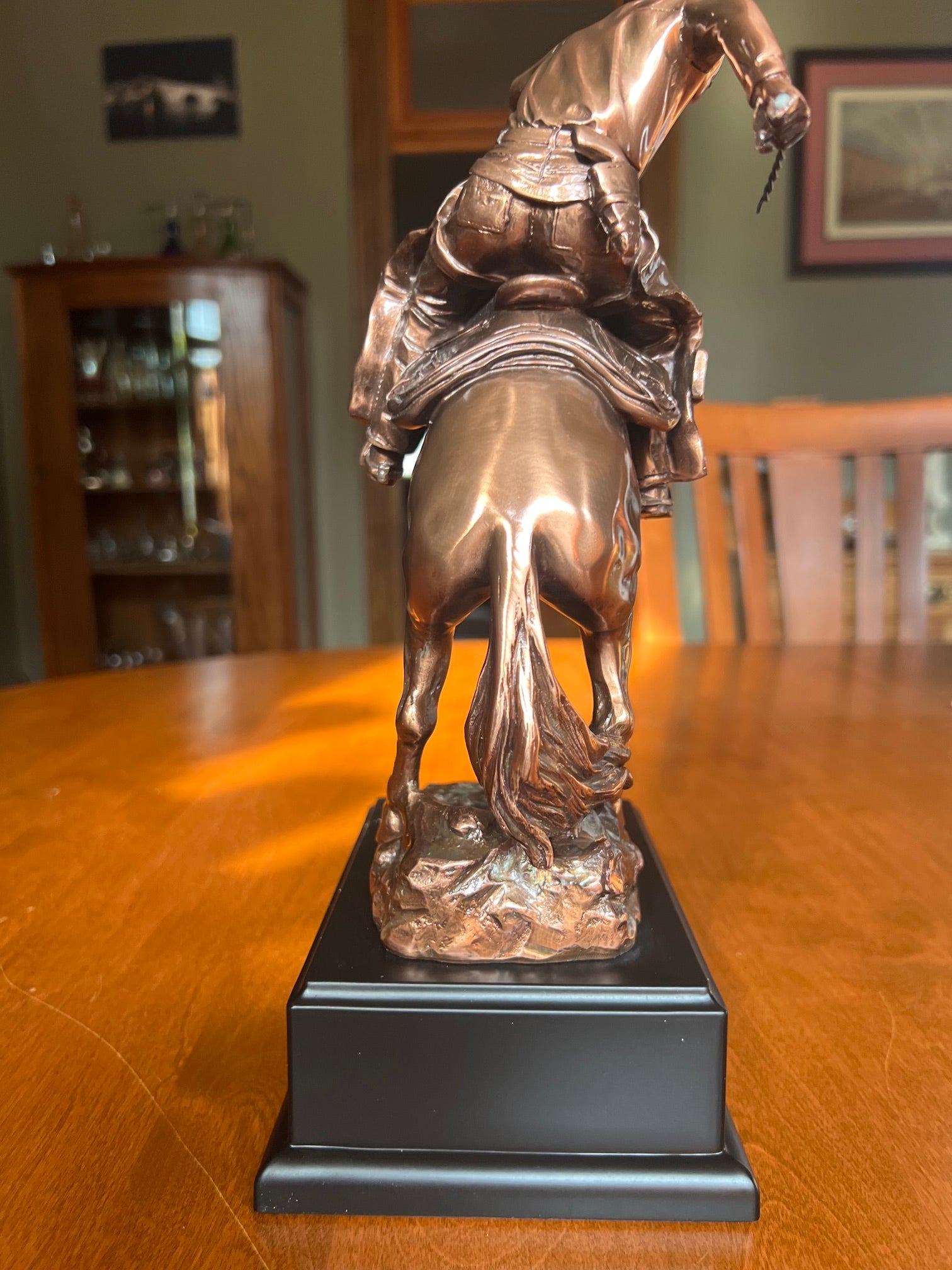 A bronze-finished sculpture of a cowboy riding a horse, mounted on a black rectangular base, displayed on a wooden table back view