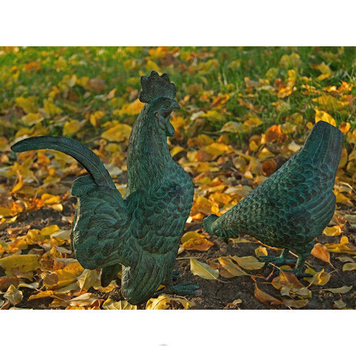 Two green garden statues of a rooster and hen placed on a ground covered with yellow autumn leaves.