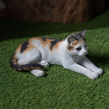Calico cat figurine lying on grass featuring detailed craftsmanship and lifelike charm