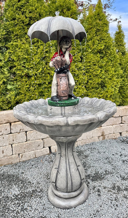 Statue of a golfer person holding an umbrella over a bird bath with greenery in the background