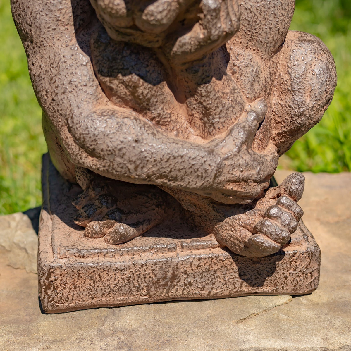 A close-up of a textured stone garden statue depicting a seated figure, shown outdoors on a stone surface with green grass in the background.