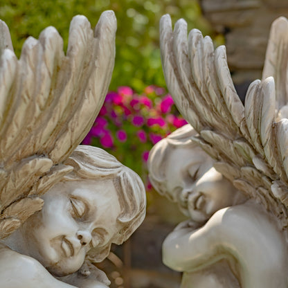 Two white angel garden statues with detailed wings and serene faces, set against a blurred background of green foliage and pink flowers.