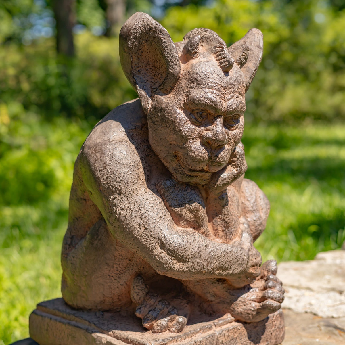 A small stone gargoyle garden statue with textured weathered brown and gray stone, shown outdoors on a stone surface with a blurred green garden background.