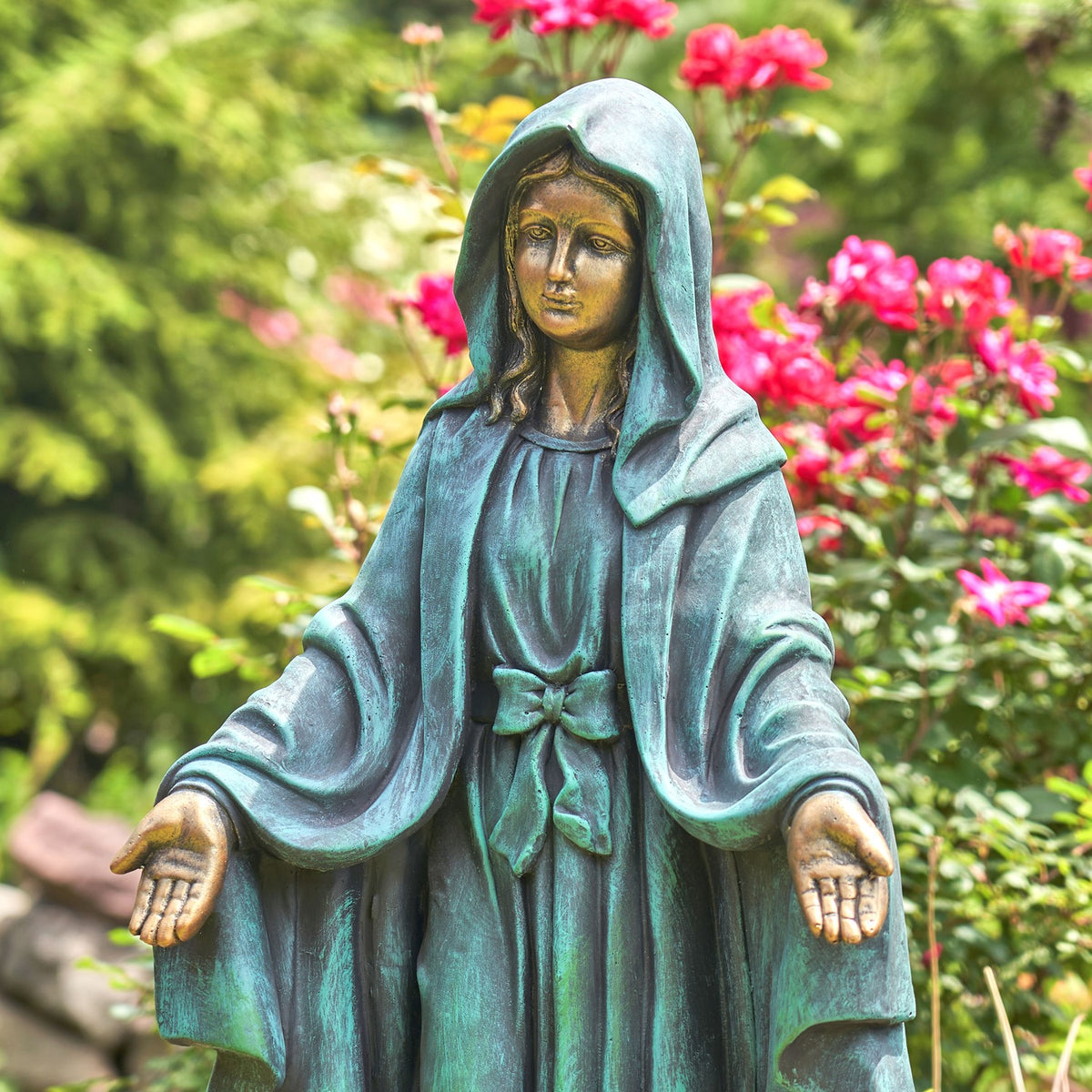 A green patina-style statue of the Virgin Mary with outstretched hands, set against a blurred garden background of green foliage and pink flowers.