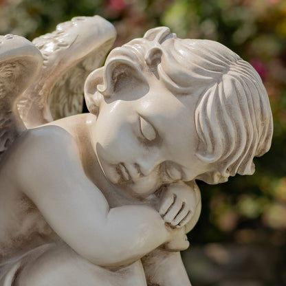 A close-up of a white stone angel garden statue with a sleeping cherub, featuring detailed wings and hair, set against a blurred green garden background.