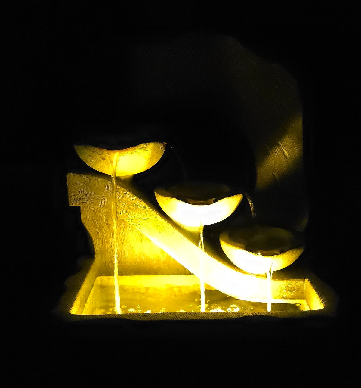 A glowing stone waterfall fountain with three illuminated bowls from which water cascades into a rectangular base, photographed against a black background.