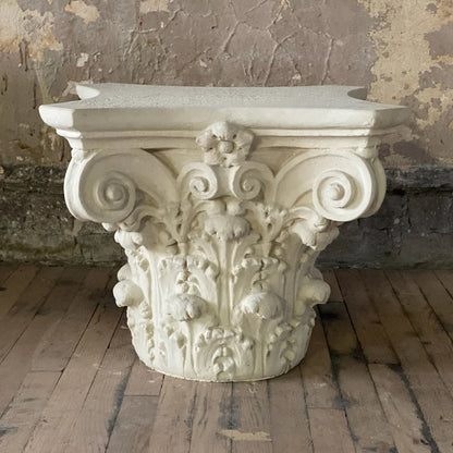 Decorative stone column capital on a wooden floor with a rustic wall background