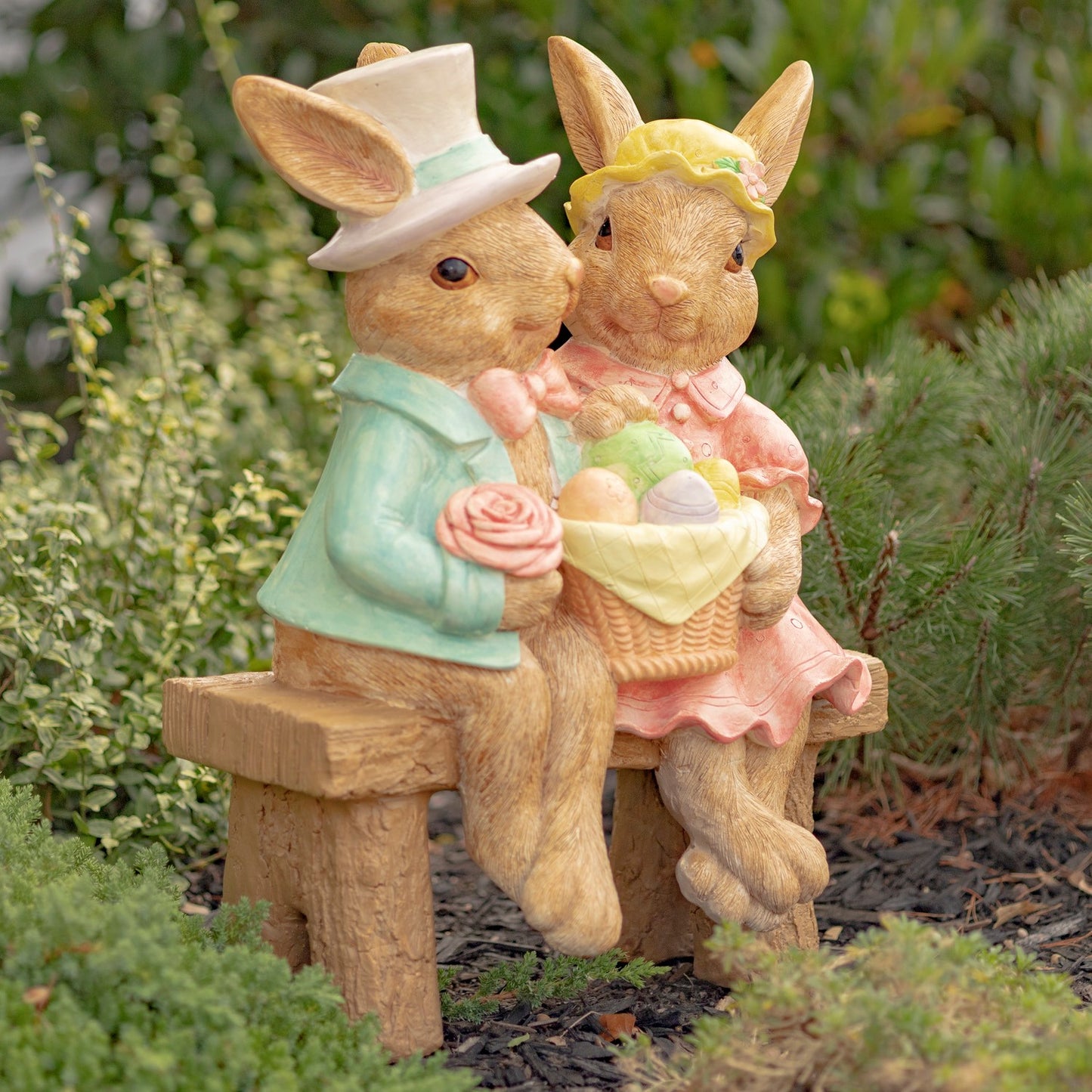 Two decorative rabbit figurines sitting on a bench with a basket of Easter eggs, wearing hats and formal attire, against a natural background.