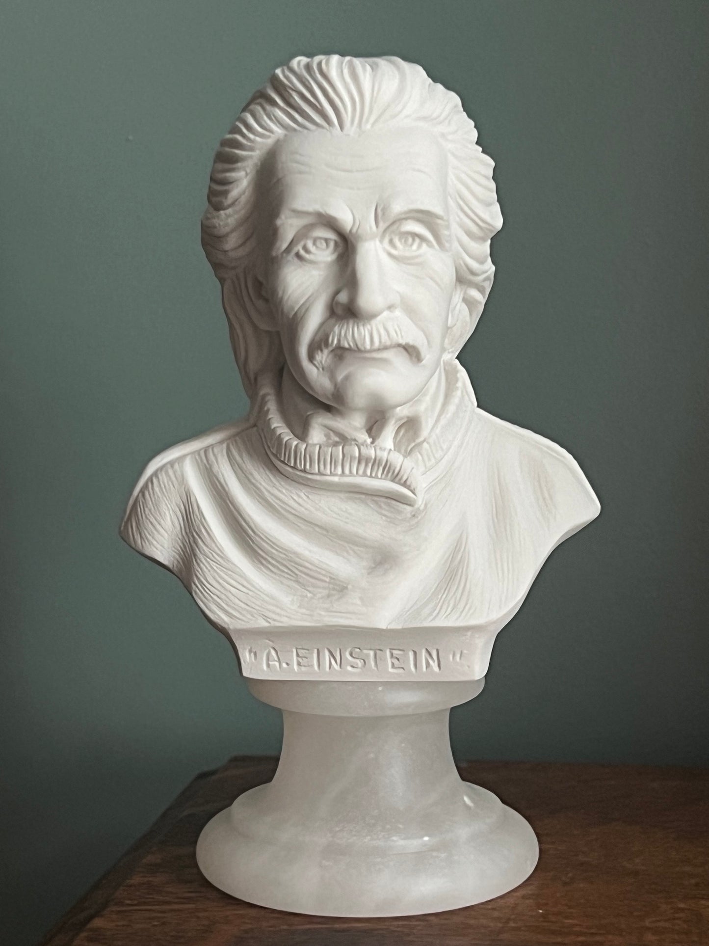 A white marble-style bust sculpture of Albert Einstein mounted on a pedestal base, photographed indoors against a muted gray wall with a dark wooden surface visible on the right.