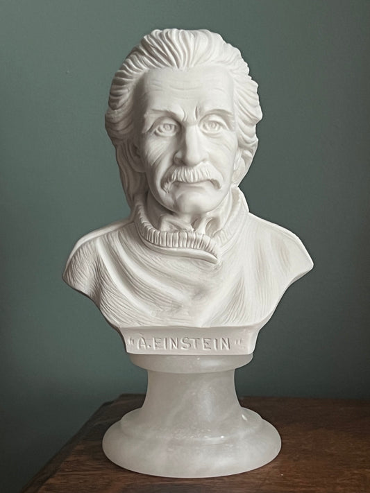 A white marble-style bust sculpture of Albert Einstein mounted on a pedestal base, photographed indoors against a muted gray wall with a dark wooden surface visible on the right.