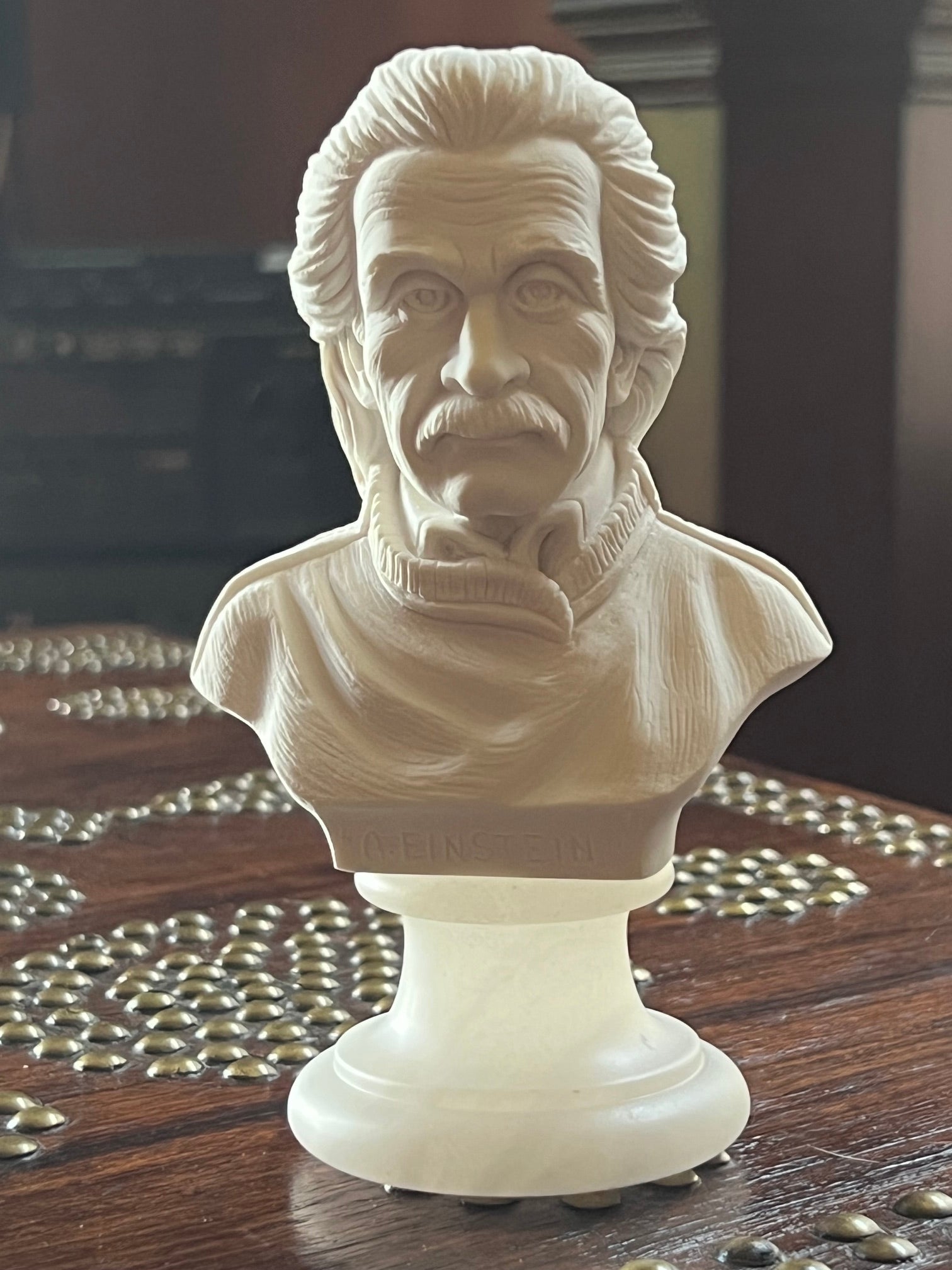 A white marble-style bust sculpture of Albert Einstein on a pedestal base, photographed indoors on a dark wooden surface with decorative metallic beads nearby.