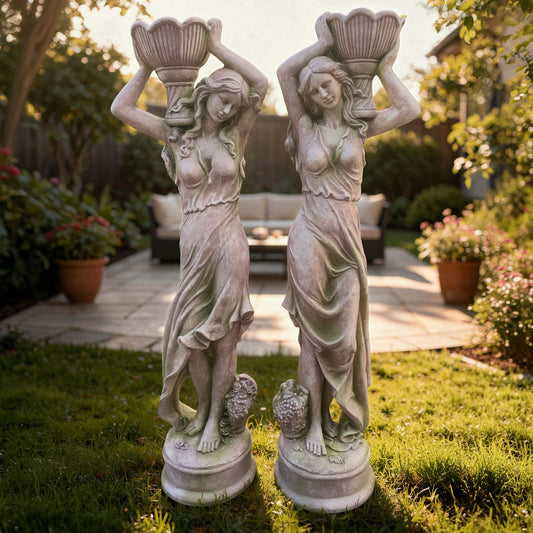 Two stone like ladies statues of women holding shell-like planters objects in a garden setting.