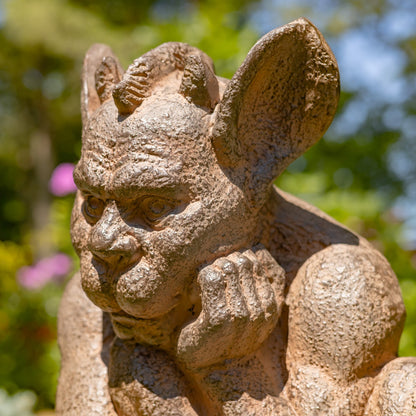 A close-up of a textured stone gargoyle garden statue with large ears and a thoughtful expression, set against a blurred green garden background.