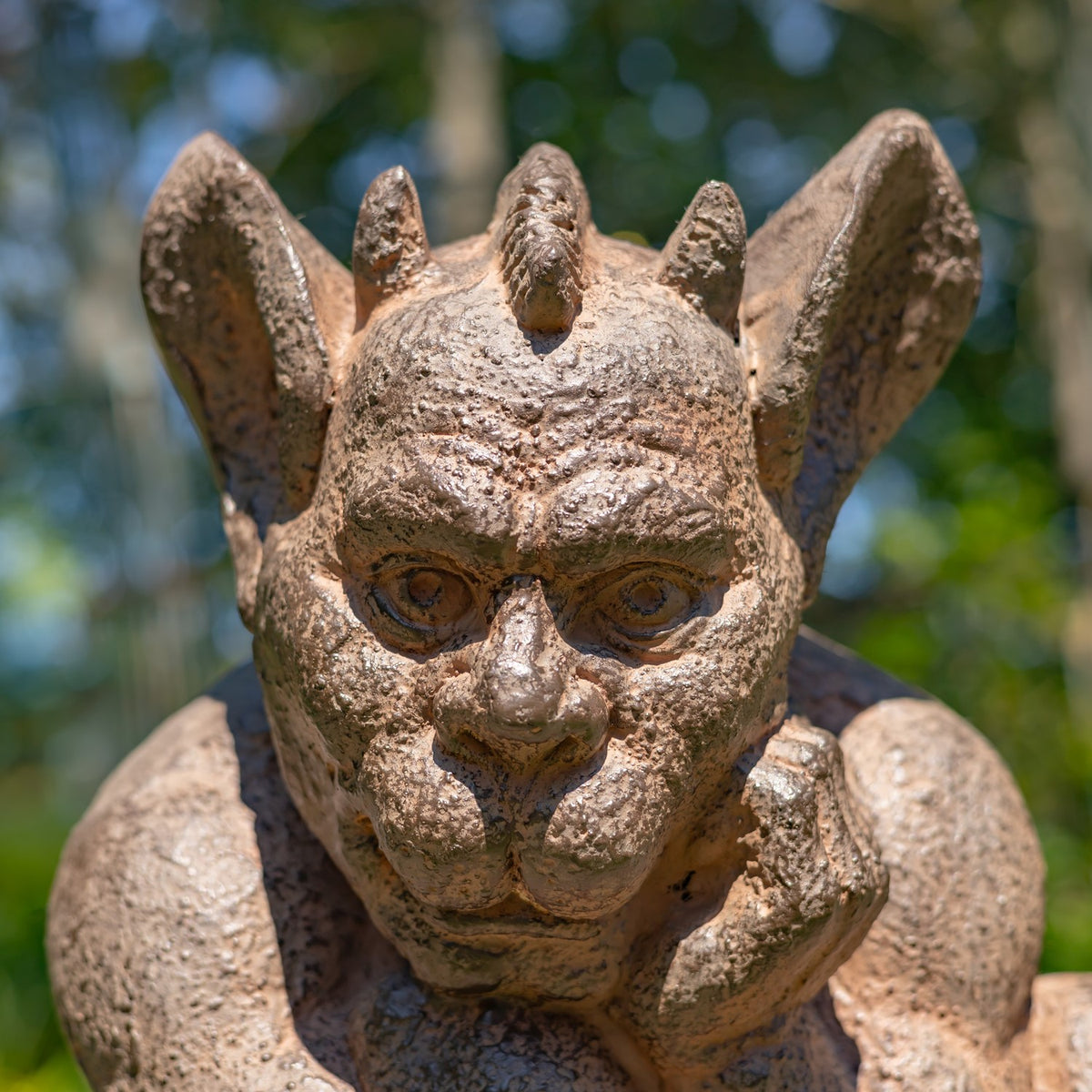 A close-up of a stone gargoyle statue with large pointed ears, a wrinkled face, and a thoughtful pose, photographed outdoors against a blurred green foliage background.