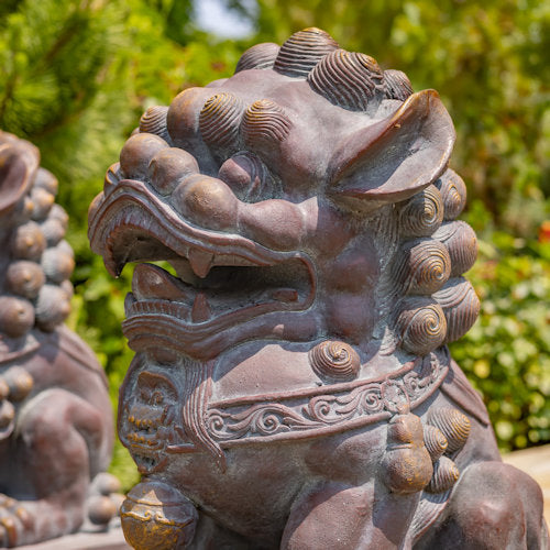 Bronze lion statue with detailed craftsmanship against a green foliage background close up of face