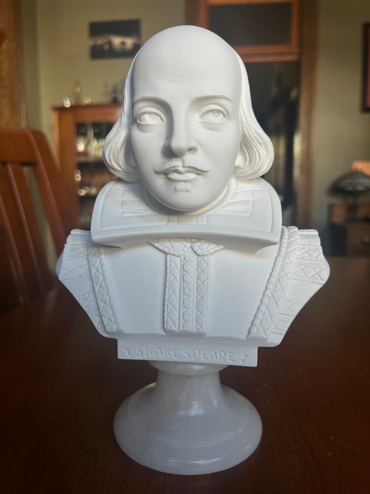 A white marble-style bust sculpture of William Shakespeare on a pedestal stand, photographed indoors on a dark wooden surface.