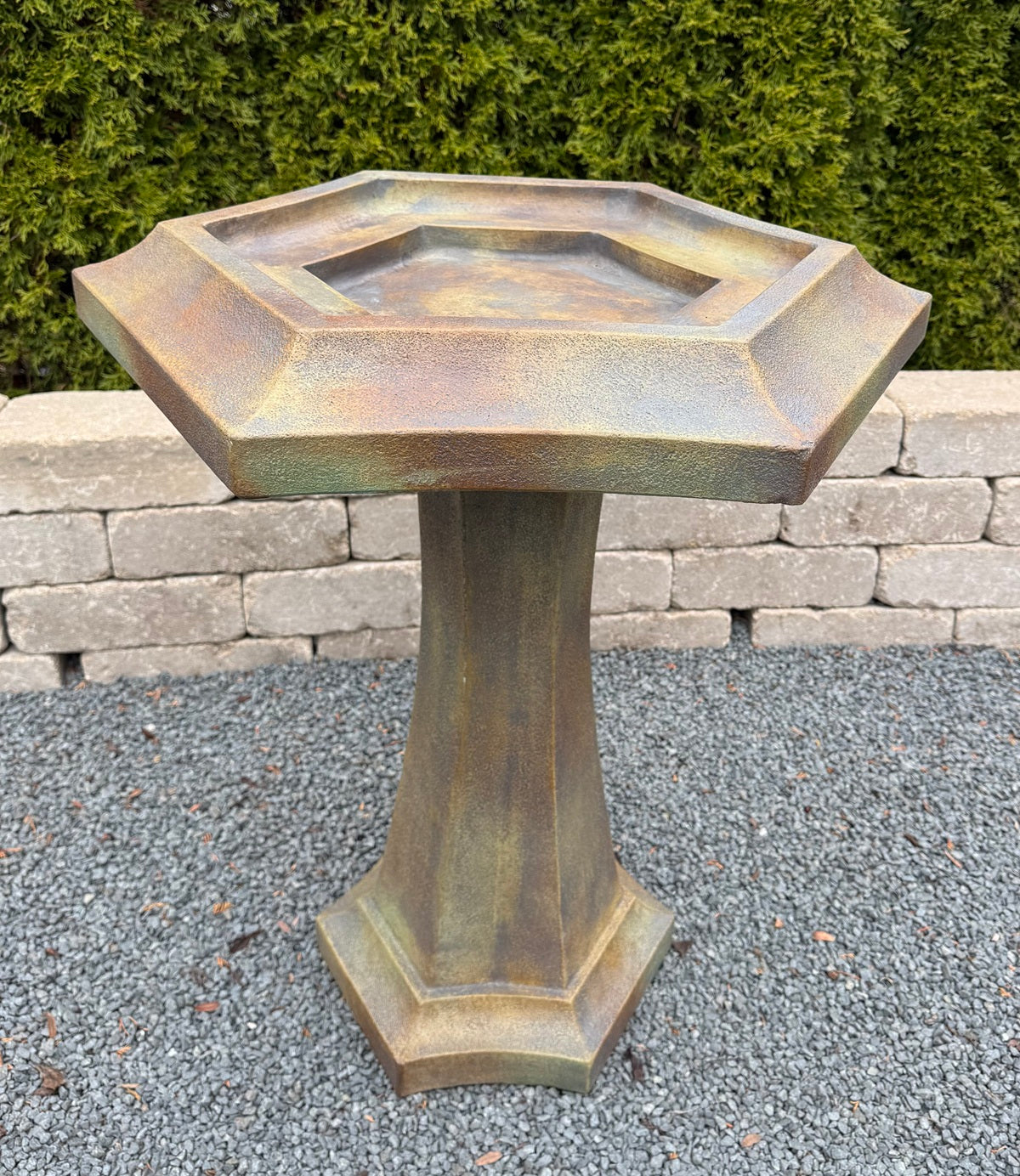 A hexagonal pedestal bird bath with a rustic bronze and brown textured finish, set outdoors on a gravel surface with a stone wall and green hedge in the background.
