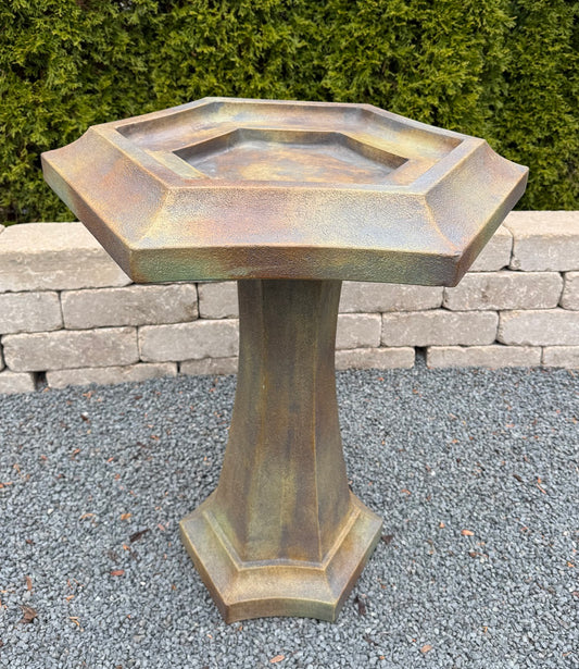 A hexagonal pedestal bird bath with a rustic bronze and brown textured finish, set outdoors on a gravel surface with a stone wall and green hedge in the background.