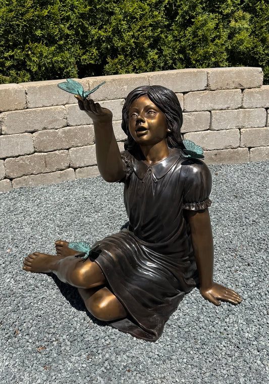 Bronze statue of a girl sitting on the ground with a butterfly near her hand, set against a stone wall and greenery.