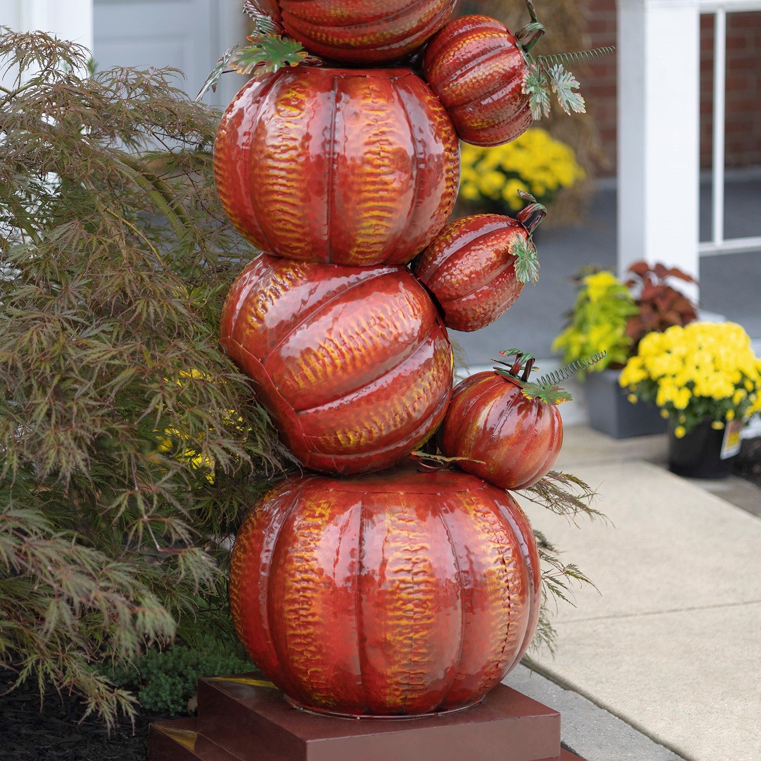 A decorative stack of glossy red and orange pumpkins with yellow-orange ribbed patterns and small green leafy tops, displayed outdoors on a pedestal in front of a house with plants and yellow flowers.