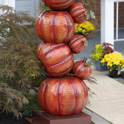 A decorative stack of glossy red and orange pumpkins with yellow-orange ribbed patterns and small green leafy tops, displayed outdoors on a pedestal in front of a house with plants and yellow flowers.
